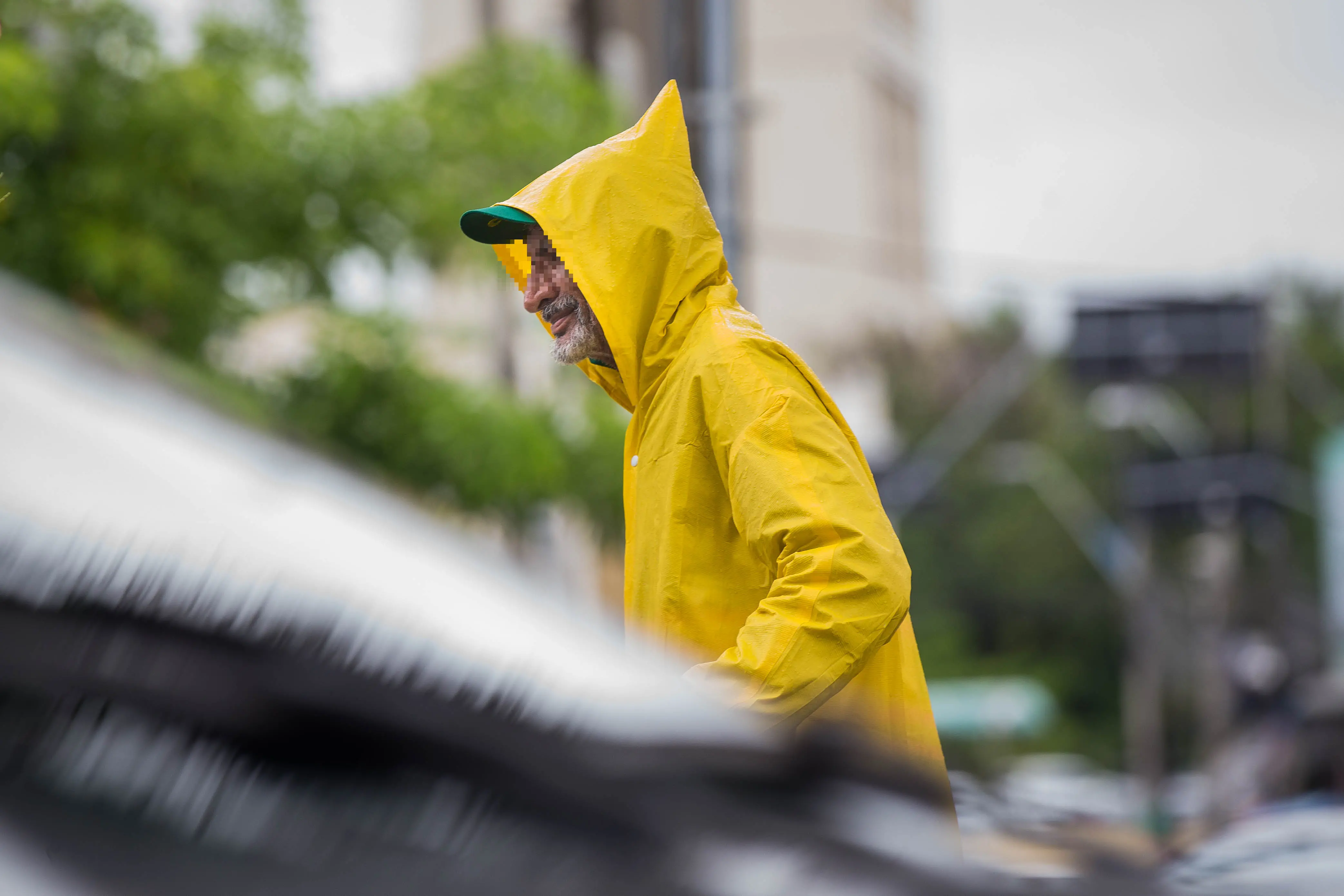Homem de barba grisalha e casaco de chuva amarelo vibrante, com um boné verde por baixo do capuz, sorrindo em um dia chuvoso.