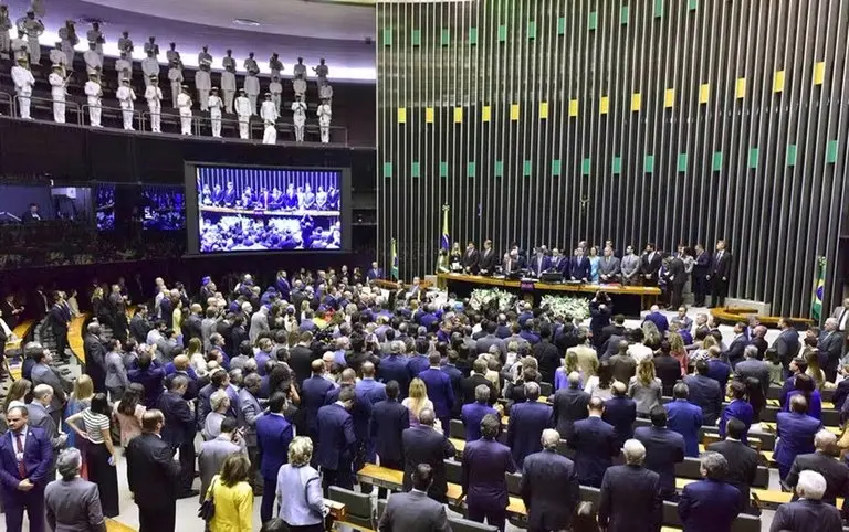 Plenário da Câmara dos Deputados lotado, com parlamentares e autoridades em pé durante uma sessão solene. Ao fundo, o painel verde e amarelo do plenário e telões exibindo a cerimônia.