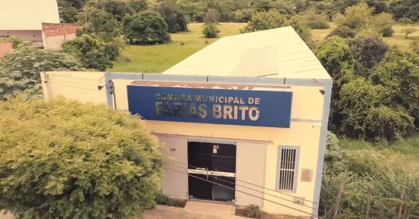 Fachada da Câmara Municipal de Farias Brito, prédio claro com placa azul com o nome da instituição, vista aérea, cercado por árvores e área verde.