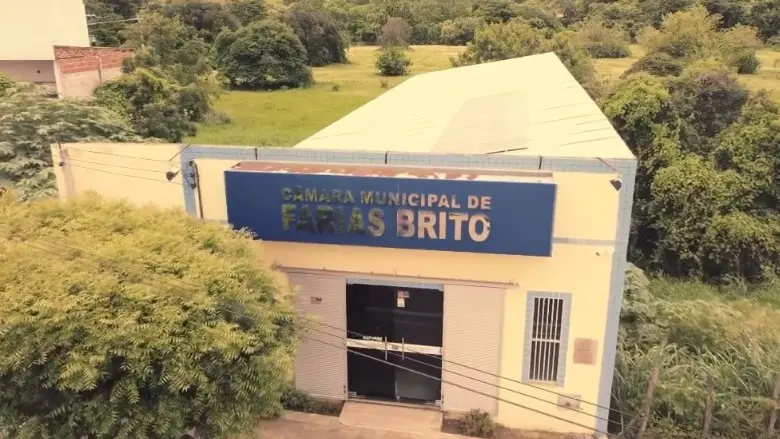 Fachada da Câmara Municipal de Farias Brito, prédio claro com placa azul com o nome da instituição, vista aérea, cercado por árvores e área verde.