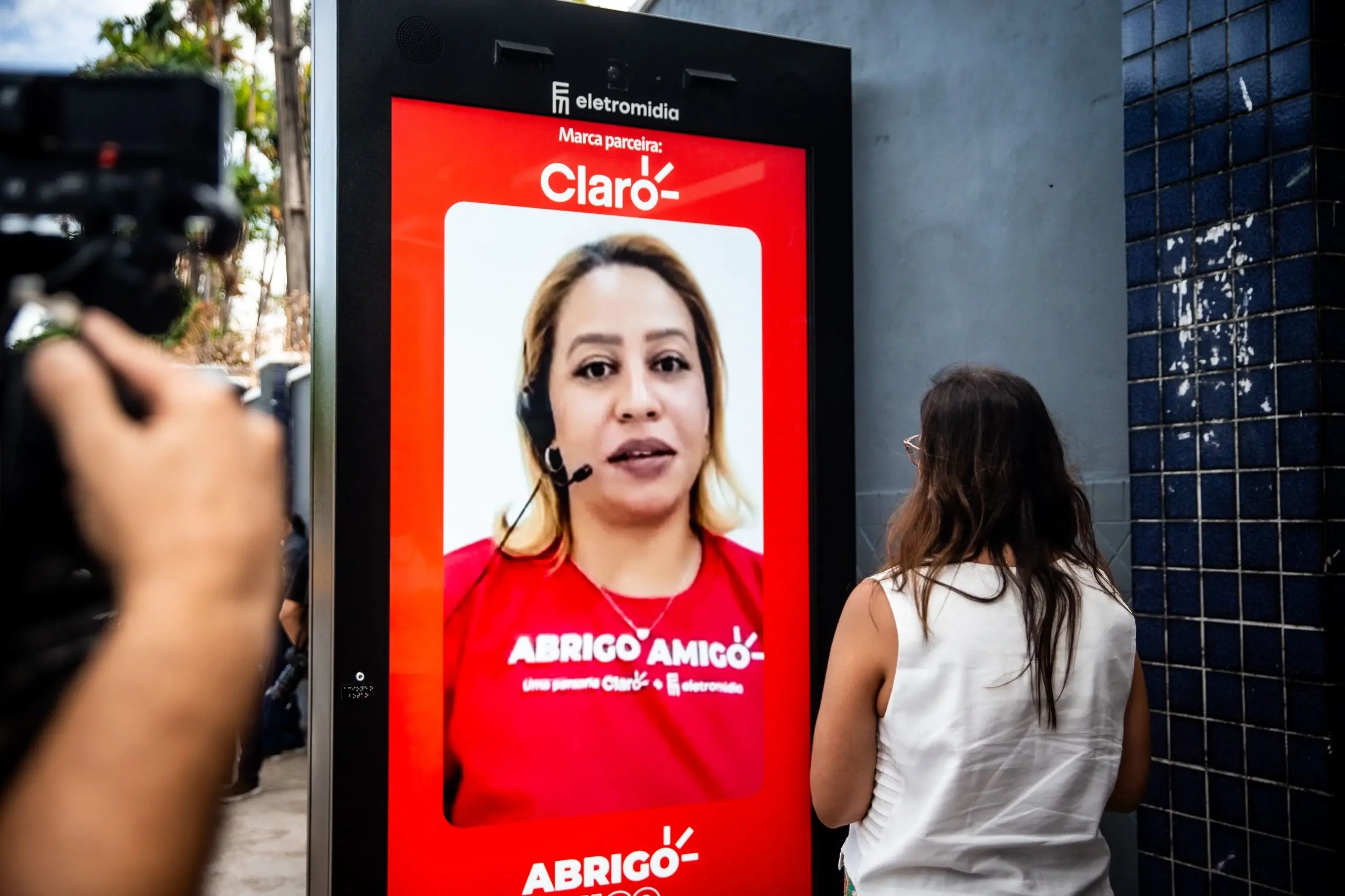 Quiosque digital da Eletromidia e Claro mostrando uma assistente virtual, enquanto uma pessoa assiste e uma câmera registra a cena.