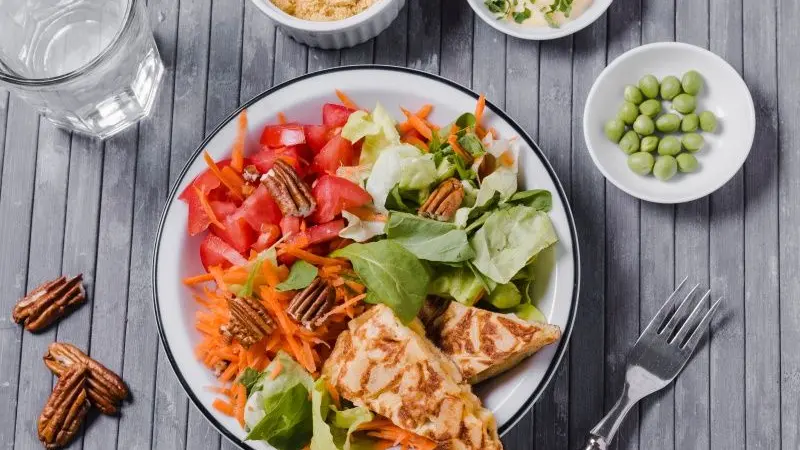 Prato de salada colorida com frango grelhado, tomate, alface, cenoura e nozes-pecã, acompanhado de água e ingredientes adicionais sobre mesa de madeira.