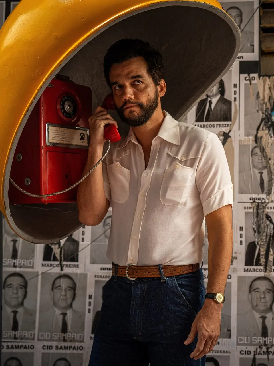 Homem usando camisa social clara e calça jeans escuras fala em um telefone público vermelho instalado dentro de uma antiga cabine amarela de rua. Ele segura o fone com uma mão enquanto a outra está no bolso. No fundo, há um muro coberto por cartazes políticos em preto‑e‑branco.