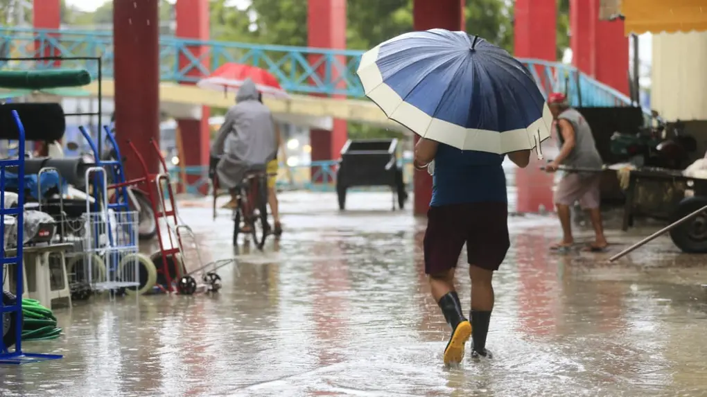 Pessoa com guarda-chuva andando na rua encharcada devido à forte chuva, durante dia nublado em uma área urbana com alguns pedestres e bicicletas ao fundo.