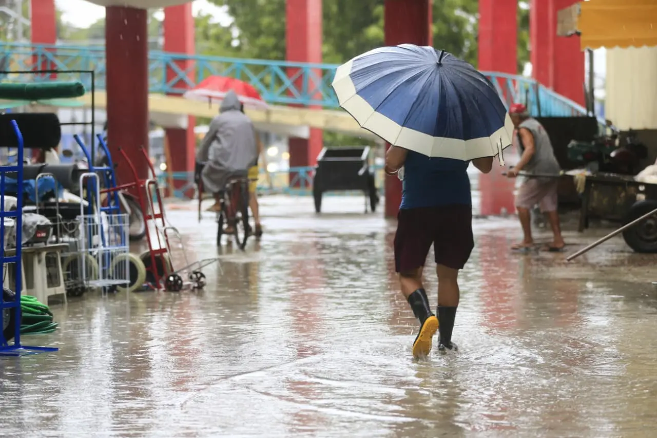 Pessoa com guarda-chuva andando na rua encharcada devido à forte chuva, durante dia nublado em uma área urbana com alguns pedestres e bicicletas ao fundo.