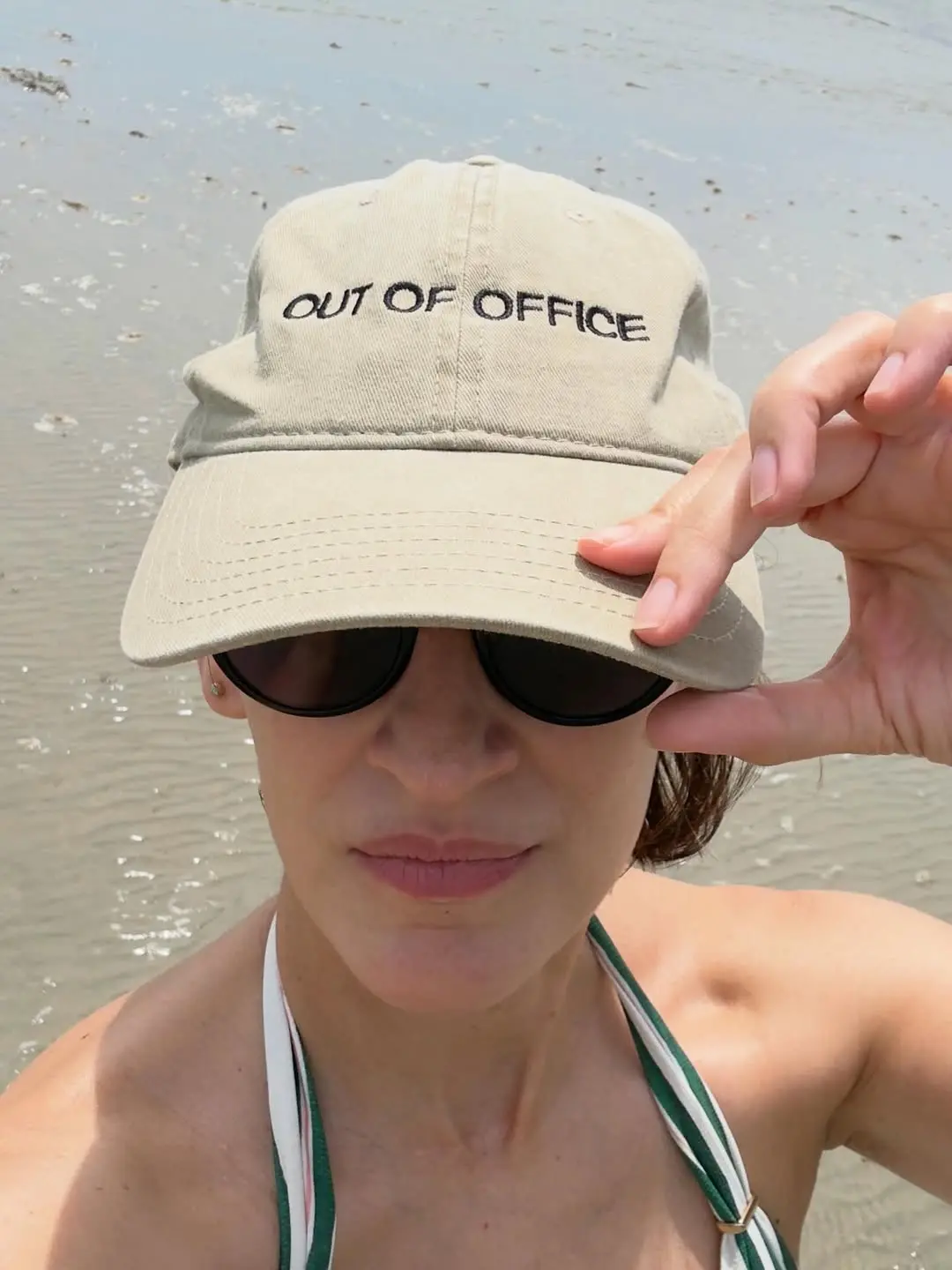 jornalista maria prata, na praia, segurando um boné com uma das mãos. no boné se lê out of office.