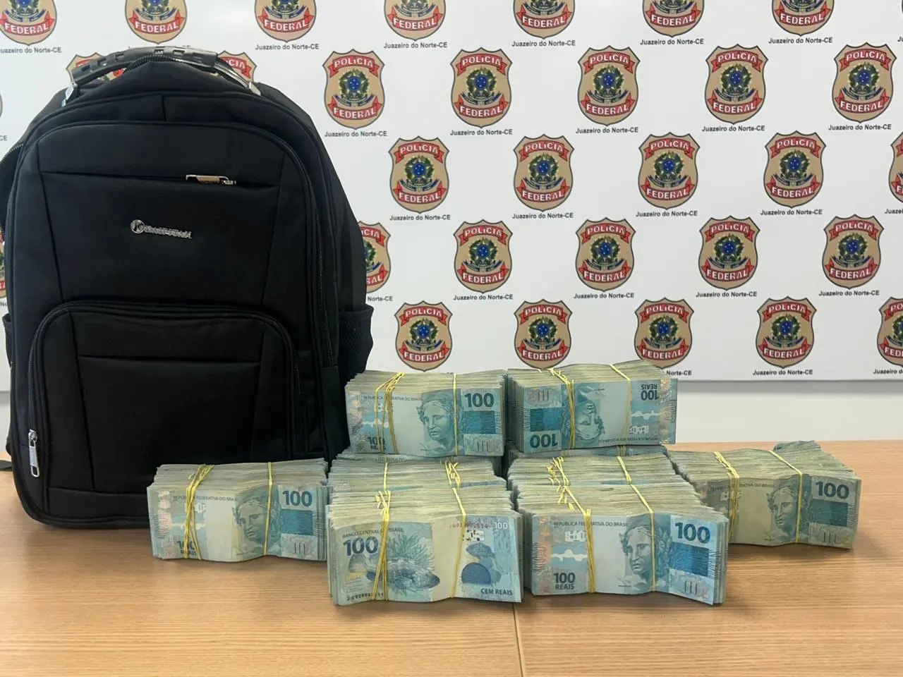 A foto mostra milhares de cédulas de R$ 100 e uma mochila em cima de uma mesa. Ao fundo, há um toldo com emblemas da Polícia Federal.