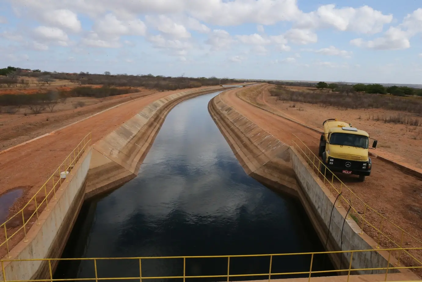 Um longo canal de irrigação do Eixão das Águas, no Ceará, de concreto com água calma atravessa uma paisagem árida de terra vermelha, com um caminhão amarelo estacionado à direita e vegetação seca ao redor.