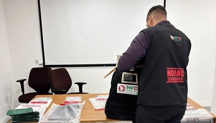 A imagem mostra um homem de roupa escura, de costas, com um colete com a inscrição Nuinc, segurando uma bolsa com a inscrição MPCE, apoiada em uma mesa, que também contém documentos.