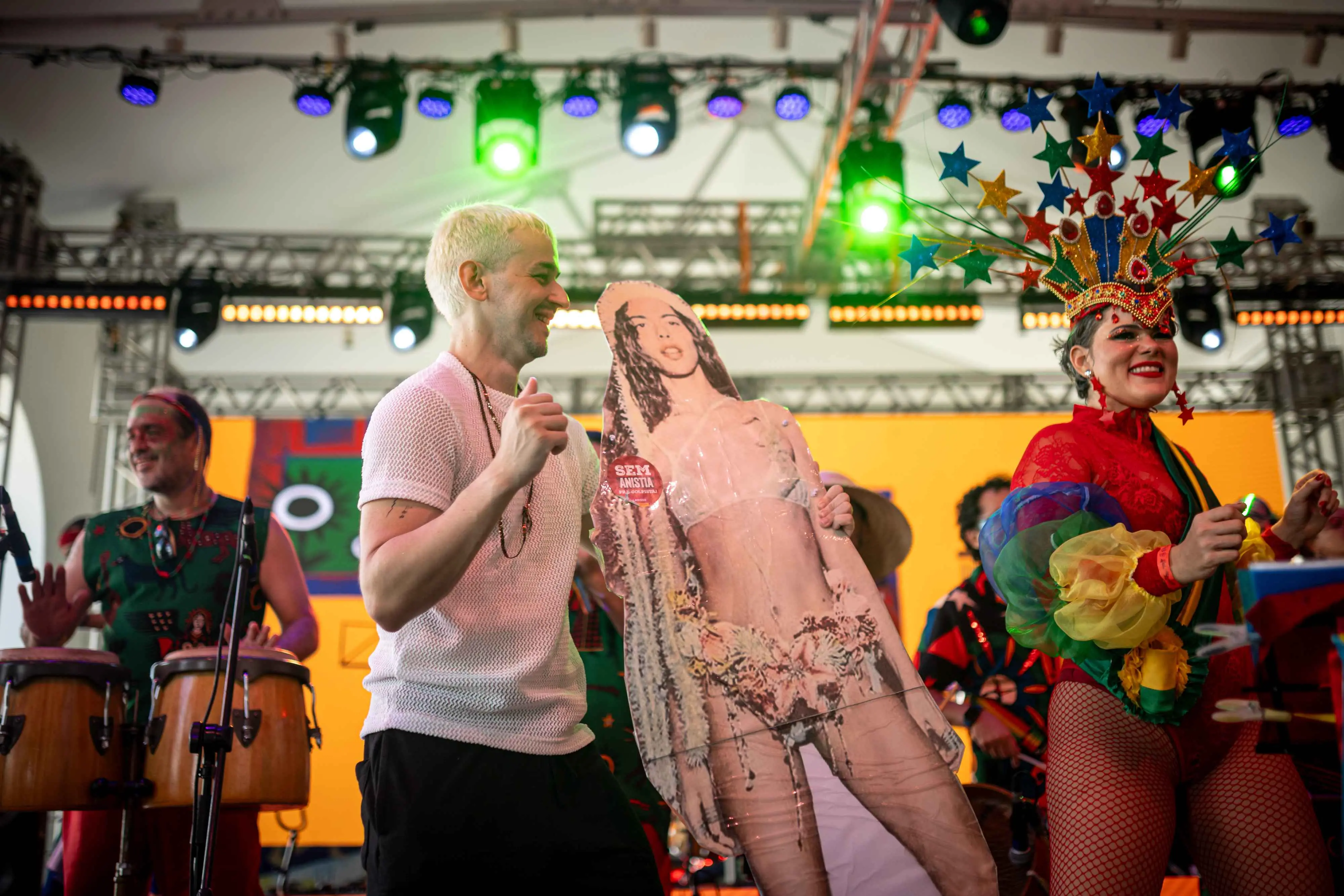Em um palco iluminado, o dançarino Juliano Floss, loiro e de camiseta branca, sorri ao segurar um totem de papelão da cantora Marina Sena. Ao lado, uma artista usa um figurino carnavalesco vibrante com um grande adorno de estrelas coloridas na cabeça. No fundo, músicos tocam percussão sob luzes de palco verdes e azuis.