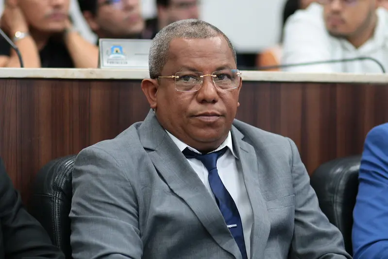 Luiz Paupina, vereador de Fortaleza, sentado em plenário, usando terno cinza, camisa branca e gravata azul, com expressão séria, em ambiente institucional com outras pessoas ao fundo desfocadas.