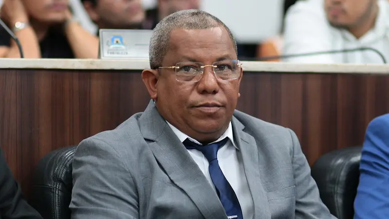 Luiz Paupina, vereador de Fortaleza, sentado em plenário, usando terno cinza, camisa branca e gravata azul, com expressão séria, em ambiente institucional com outras pessoas ao fundo desfocadas.