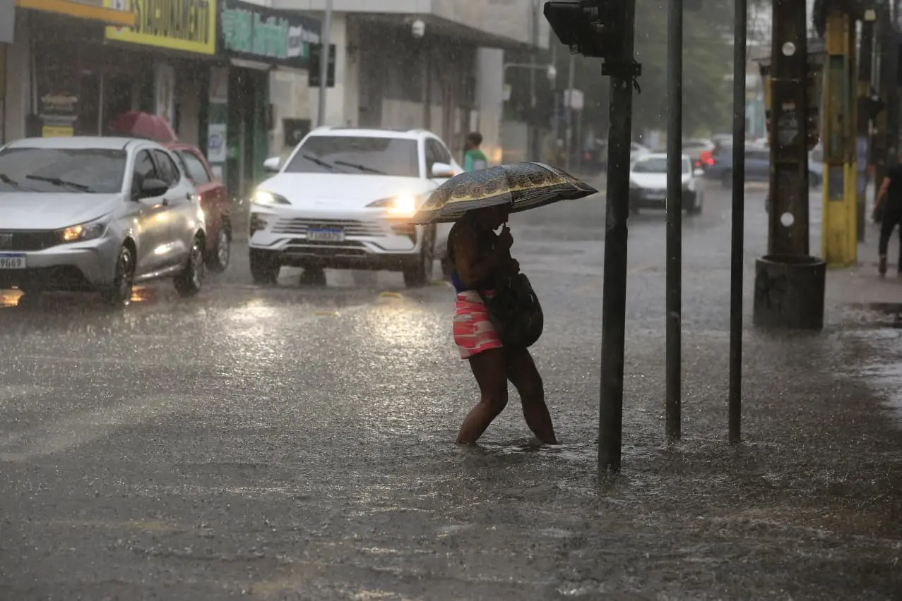 Imagem mostra uma mulher atravessando a rua alagada. Imagem usada em matéria sobre alertas de risco da Defesa Civil.