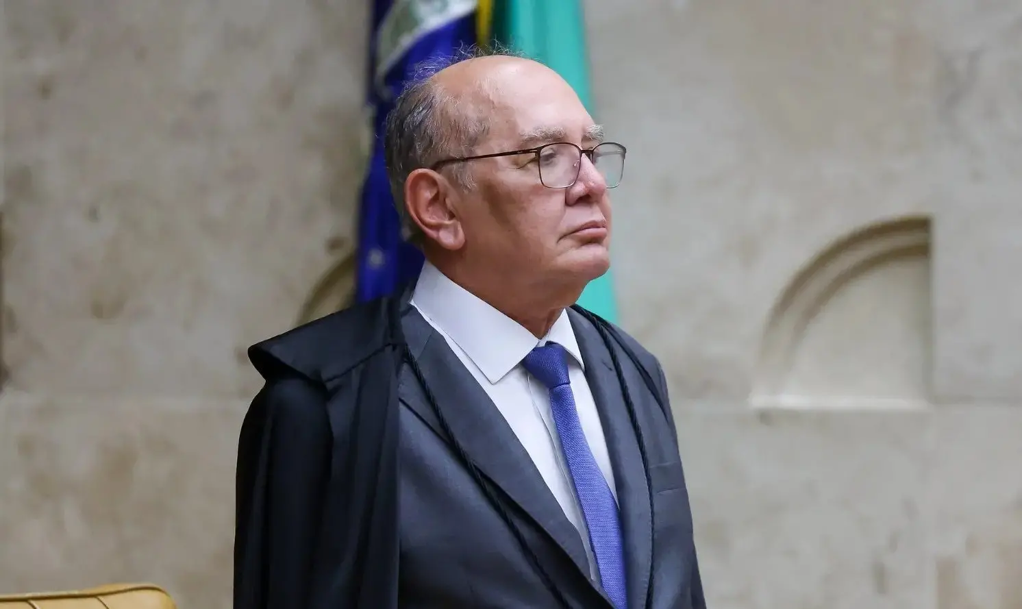 Gilmar Mendes é um homem idoso, calvo e usa óculos de grau de armação preta e fina. Na foto, ele está de lado e usa terno preto com camisa social branca e gravata azul. Imagem usada em matéria onde ministro dá 48 horas para que Santa Catarina explique a proibição de cotas.