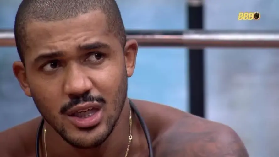 Close-up de Matheus, do BBB 26. ele é um homem jovem, negro, com cabelo raspado, bigode e barba rala, olhando para o lado direito com a boca levemente aberta, em ambiente interno.