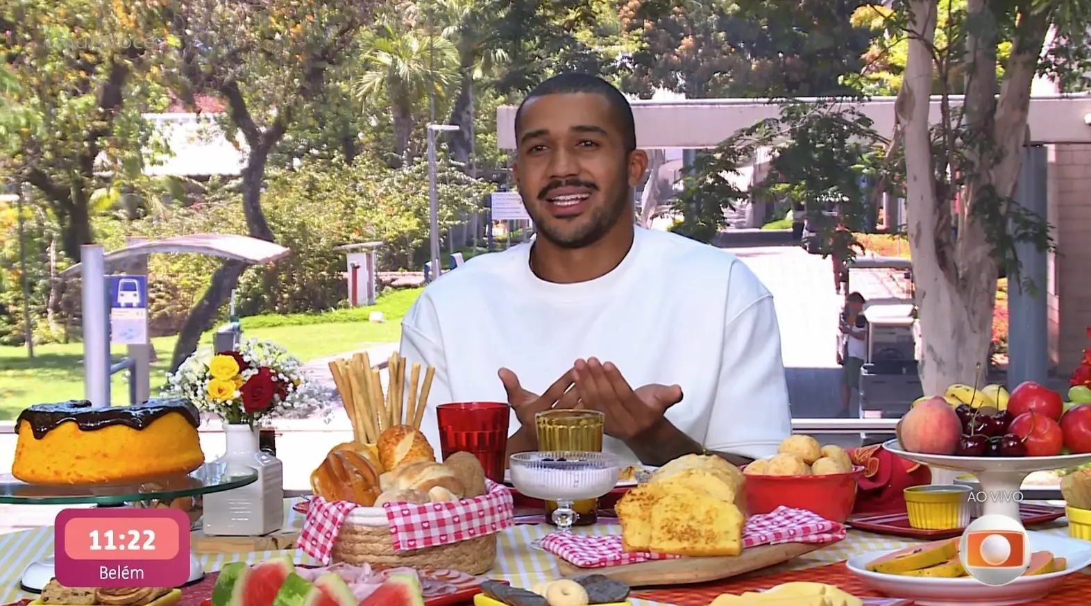 Uma mesa posta ao ar livre com diversos alimentos de café da manhã, incluindo bolo com cobertura de chocolate, pães, frutas, biscoitos, geleias e bebidas em copos coloridos. Ao fundo, há árvores, gramado e uma área urbana com pessoas passando. Uma pessoa sentada à mesa veste uma camisa branca e gesticula com as mãos enquanto participa do cenário de transmissão ao vivo, indicado por um gráfico no canto inferior direito com o logotipo da emissora.