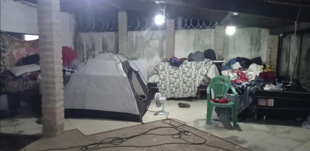 A foto mostra uma parte de um imóvel em que há barracas, camas, cadeiras e um ventilador, além de outros objetos.