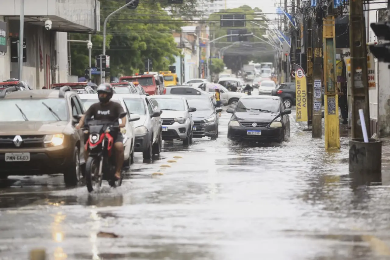 Vista de uma rua urbana alagada, com carros e uma motocicleta trafegando lentamente pela água, sob céu nublado.