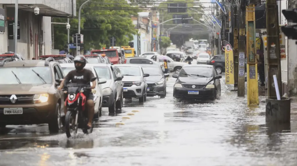 Vista de uma rua urbana alagada, com carros e uma motocicleta trafegando lentamente pela água, sob céu nublado.