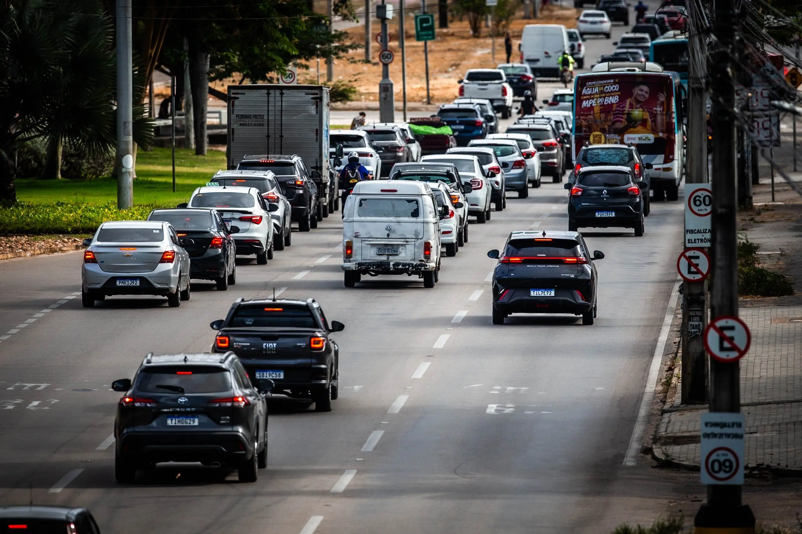 Uma avenida movimentada apresenta fluxo intenso de veículos, com carros de diversas cores e modelos distribuídos em múltiplas faixas sob luz diurna. À direita, postes exibem placas de sinalização de trânsito, como limite de velocidade de 60 km/h e proibição de estacionamento. O cenário inclui árvores à esquerda e fios elétricos suspensos que cruzam a via ao fundo.