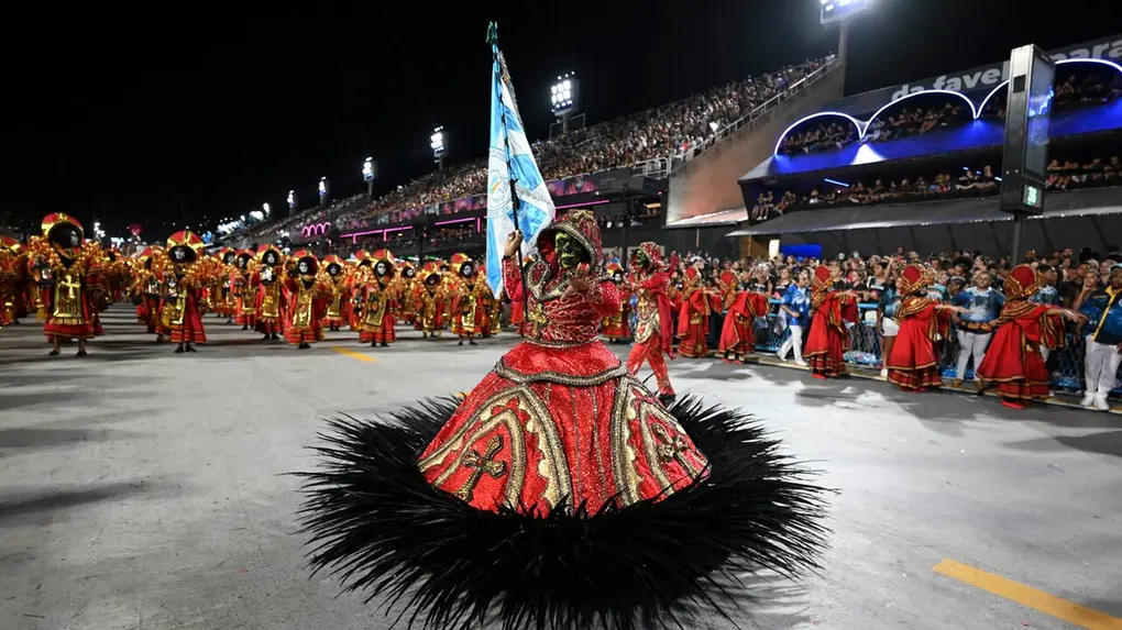 Pessoa fantasiada com vestido longo vermelho de adornos dourados e bainha preta, desfilando em rua a noite.