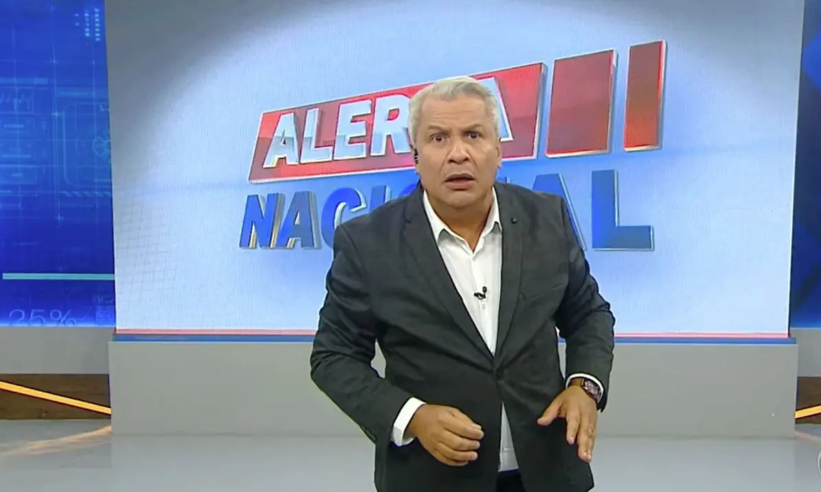 Apresentador de terno escuro e camisa branca aparece em um estúdio de televisão, em pé, com uma das mãos levantadas. Ao fundo, há um grande painel com o logotipo do programa ‘Alerta Nacional’, exibindo letras vermelhas e azuis sobre fundo claro.