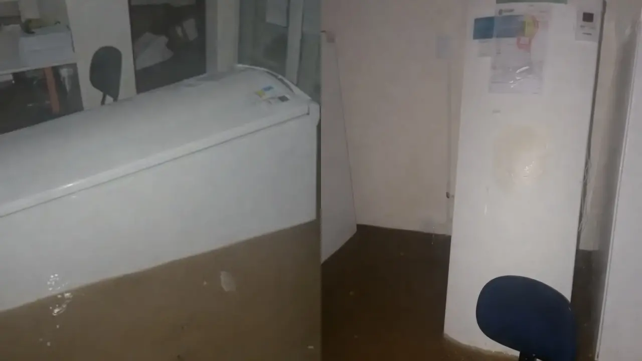 Ambiente interno alagado, com água barrenta cobrindo o piso e atingindo a base de um freezer horizontal e de uma geladeira branca; ao fundo, móveis, equipamentos e uma cadeira azul aparecem parcialmente submersos, evidenciando os impactos da inundação no local.