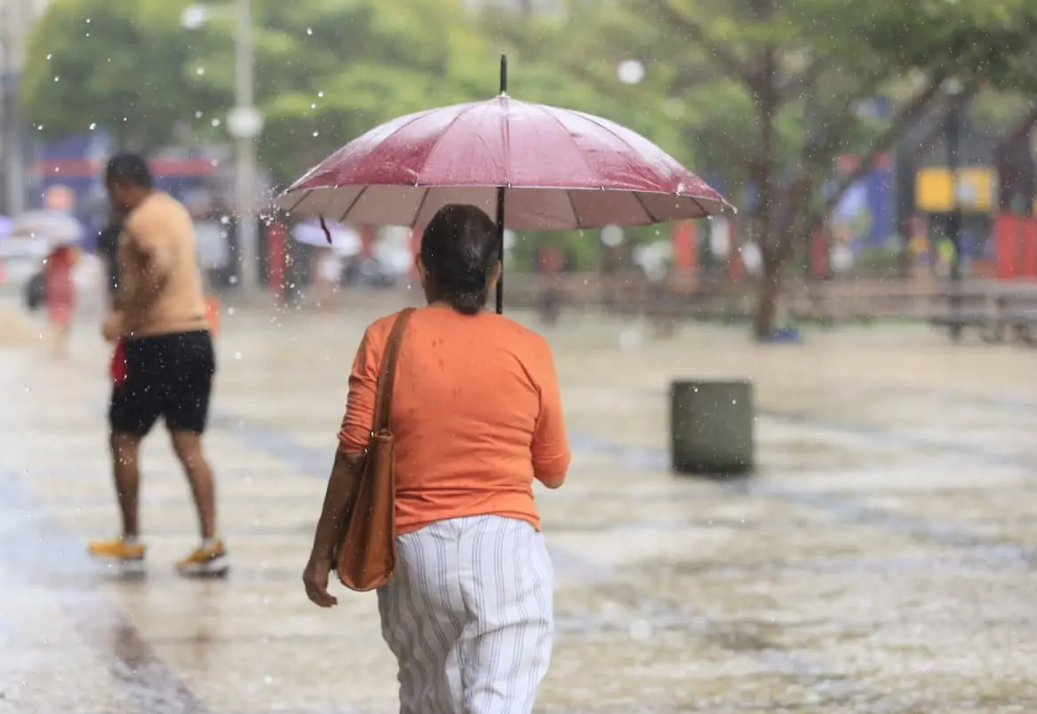 Pessoa com guarda-chuva deitada na chuva em um parque urbano com árvores e bancos ao fundo, vestindo blusa laranja e calça listrada.