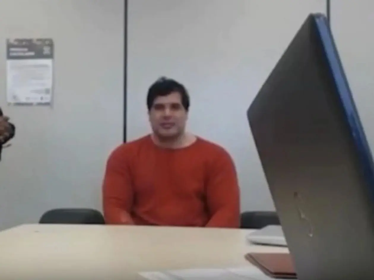 Pessoa sentada à mesa em uma sala de reuniões, vestindo uma camisa vermelha, com um laptop aberto em primeiro plano e cartaz na parede ao fundo.