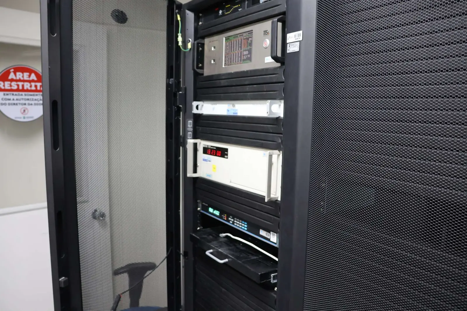 Rack de TI mostrando múltiplos equipamentos, como servidores e dispositivos de rede de computadores, em uma área de acesso controlado em Fortaleza. Ao centro, há um relógio atômico branco com letreiros vermelhos.