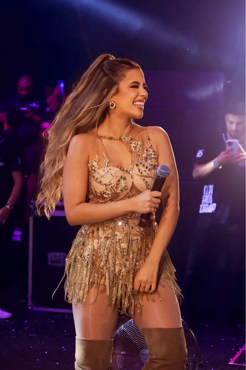 Mulher de cabelo longo e vestido com lantejoulas sorrindo e com microfone em uma das mãos.