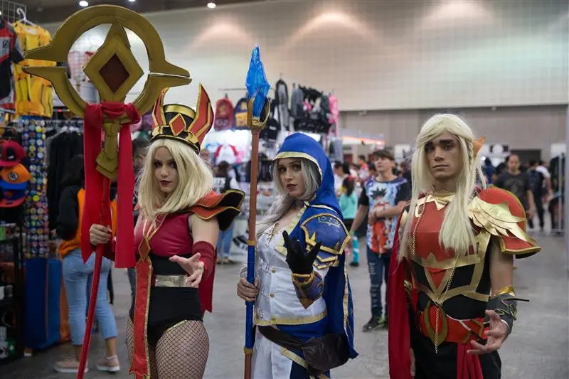 Três cosplayers posam em um pavilhão de eventos: à esquerda, uma mulher de cabelos loiros segura um grande cetro dourado; ao centro, uma jovem veste uma capa azul com capuz e segura um cajado de cristal; e à direita, um rapaz usa armadura vermelha e dourada. Eles vestem figurinos detalhados inspirados em personagens de jogos, cercados por estandes de produtos ao fundo.