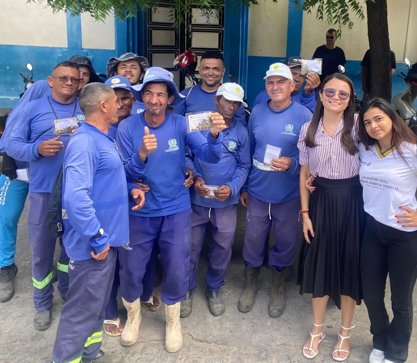 Na imagem, um grupo de aproximadamente doze pessoas posa para uma foto em frente a um prédio de fachada azul e branca. A maioria são homens vestindo uniformes de trabalho azuis (calças e blusas de manga comprida), alguns usando chapéus de aba larga ou bonés. Eles sorriem e alguns seguram pequenos pacotes plásticos transparentes contendo sementes. À direita, duas mulheres jovens sorriem; uma veste camisa listrada e saia preta, e a outra veste uma camiseta branca de uniforme escolar. O ambiente é externo, durante o dia, sob a sombra de uma árvore.