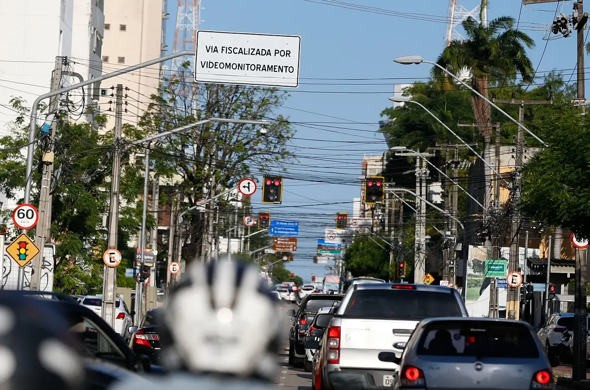 Cena urbana diurna mostrando na avenida Antonio Sales, em Fortaleza, com veículos parados no trânsito, múltiplos postes de energia com fiação e diversas placas de sinalização, incluindo o limite de velocidade (60 km/h) e videomonitoramento por câmeras.