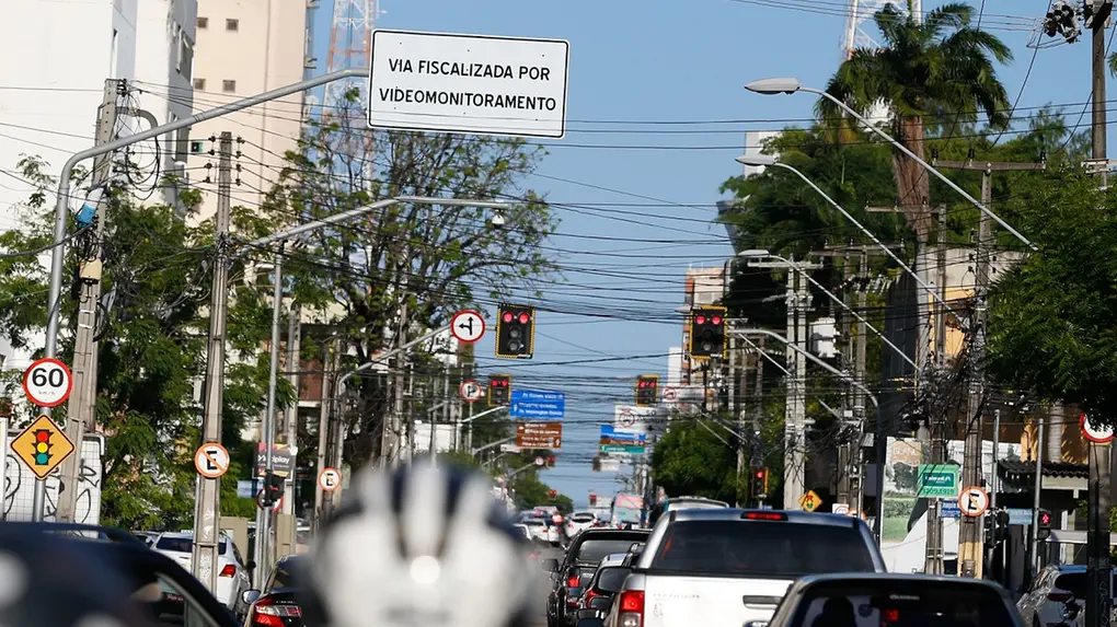 Cena urbana diurna mostrando na avenida Antonio Sales, em Fortaleza, com veículos parados no trânsito, múltiplos postes de energia com fiação e diversas placas de sinalização, incluindo o limite de velocidade (60 km/h) e videomonitoramento por câmeras.