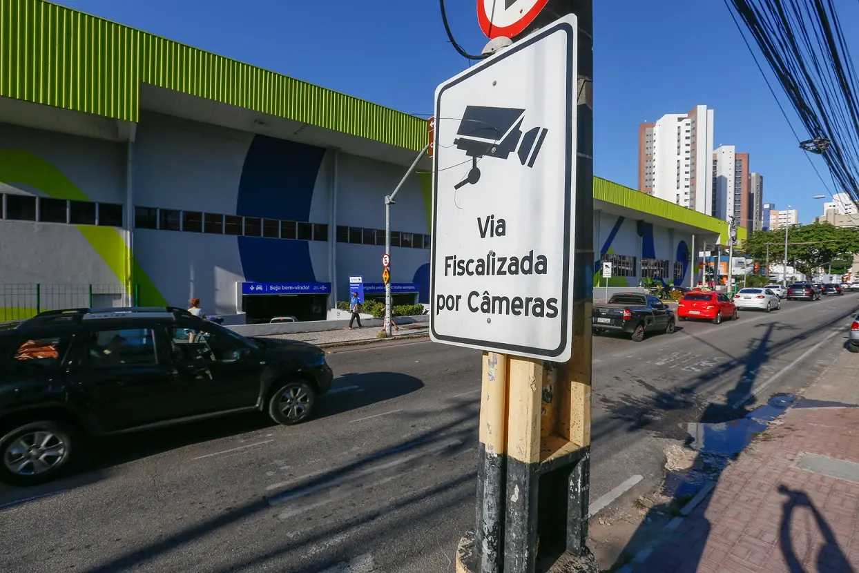 Placa de trânsito Via Fiscalizada por Câmeras em uma avenida urbana movimentada de Fortaleza, com veículos, um grande edifício e arranha-céus ao fundo.