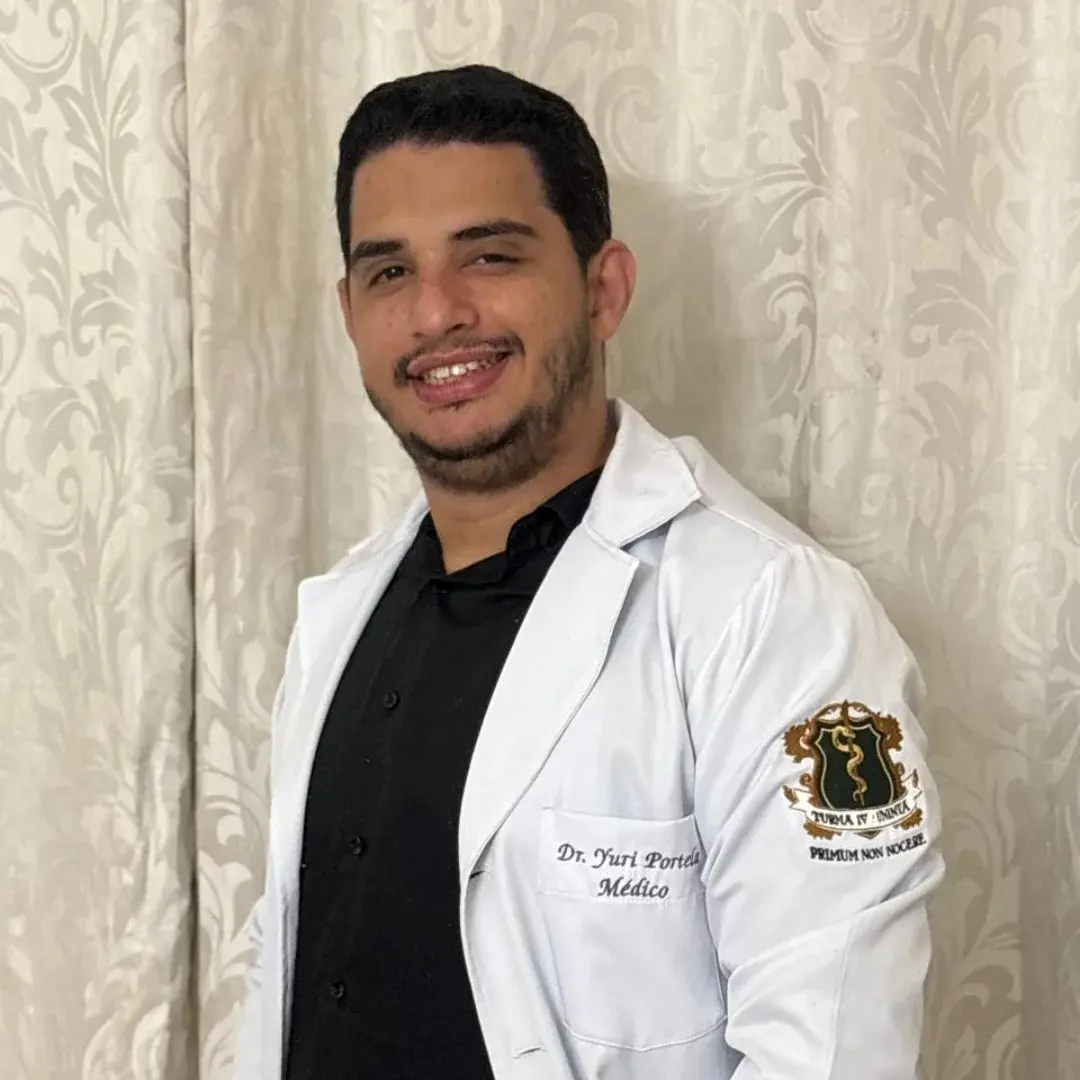 Homem com jaleco médico branco onde se lê 'Dr. Yuri Portela Médico', sorrindo para a câmera, com um emblema no braço.