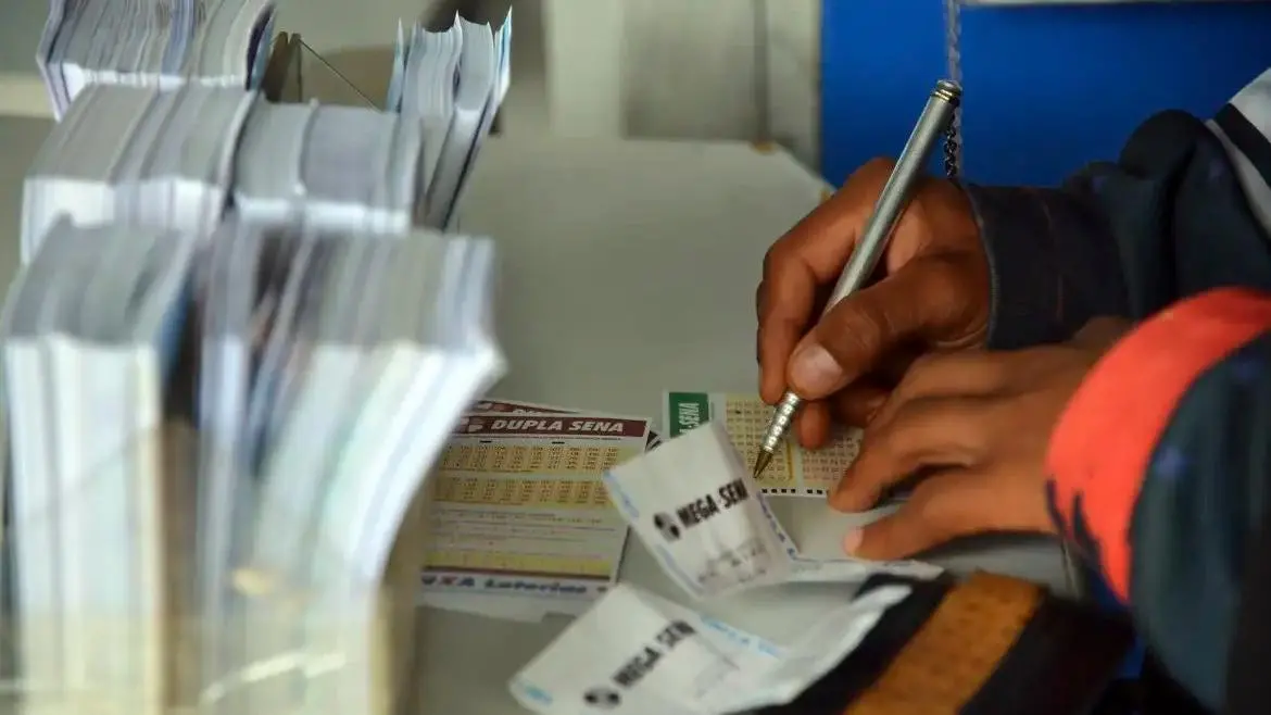 Pessoa comprando bilhetes de loteria ou raspadinhas em uma banca de jornal, com foco na mão que segura o papel e nas apostas.