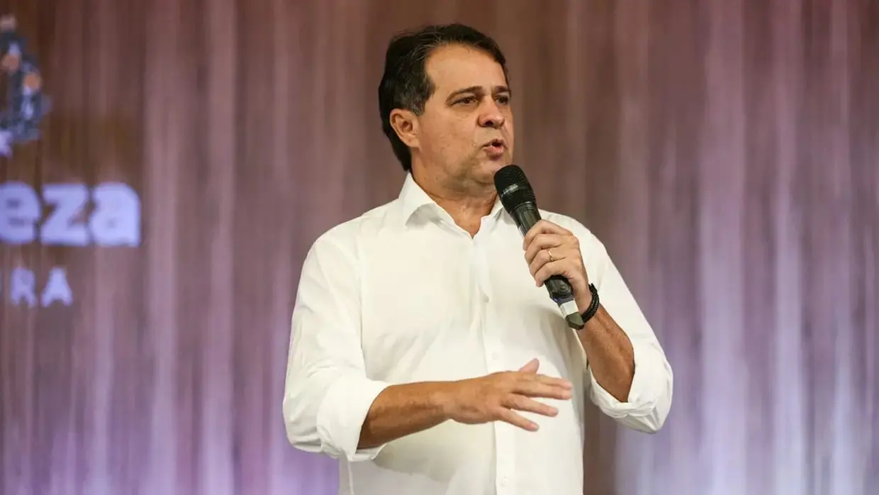 Evandro Leitão em pé, usando camisa branca de mangas longas, segura um microfone e fala em um palco, com fundo de madeira e iluminação suave, em contexto de evento ou discurso público.