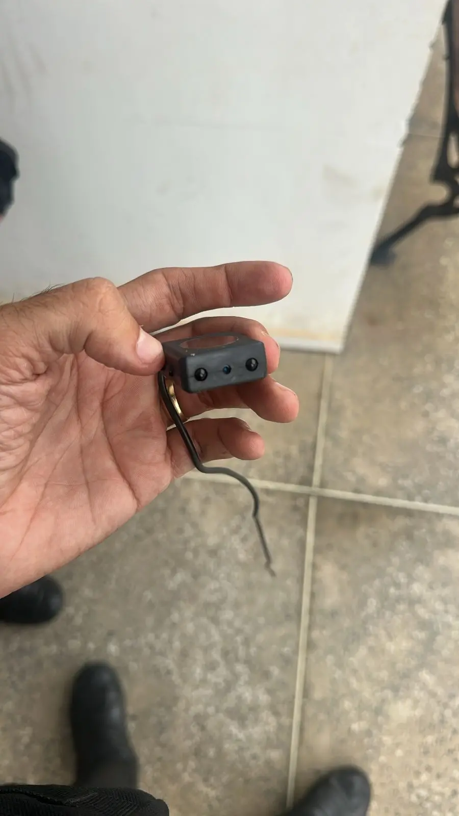 Mão de uma pessoa segurando uma pequena microcâmera preta, com fios expostos pendendo para baixo. O fundo mostra um piso de cimento e parte de uma parede clara, além de botas pretas visíveis na parte inferior da imagem.