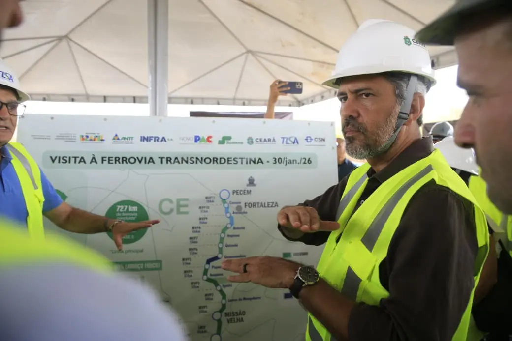 Governador Elmano de Freitas em visita ao canteiro de obras da Transnordestina. Ele veste colete de sinalização e capacete de proteção, em frente a uma imagem do mapa da obra com a atualização da execução.