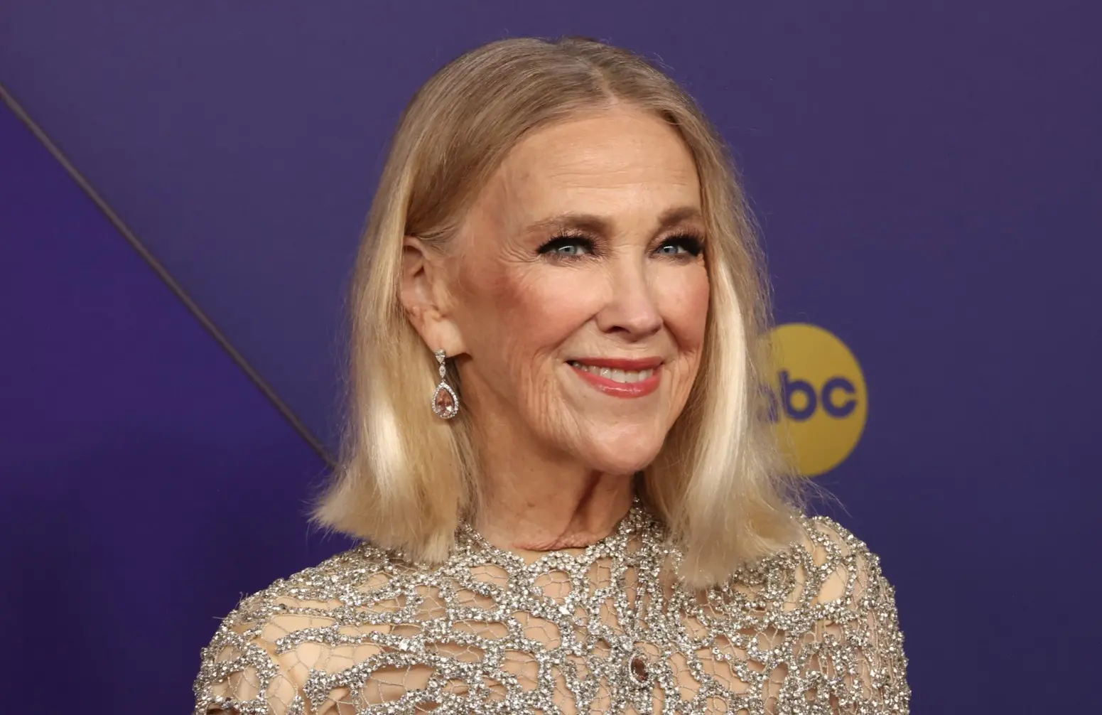 Catherine O Hara era uma mulher loira, branca e idosa. Na foto, ela usa um vestido prata brilhoso.