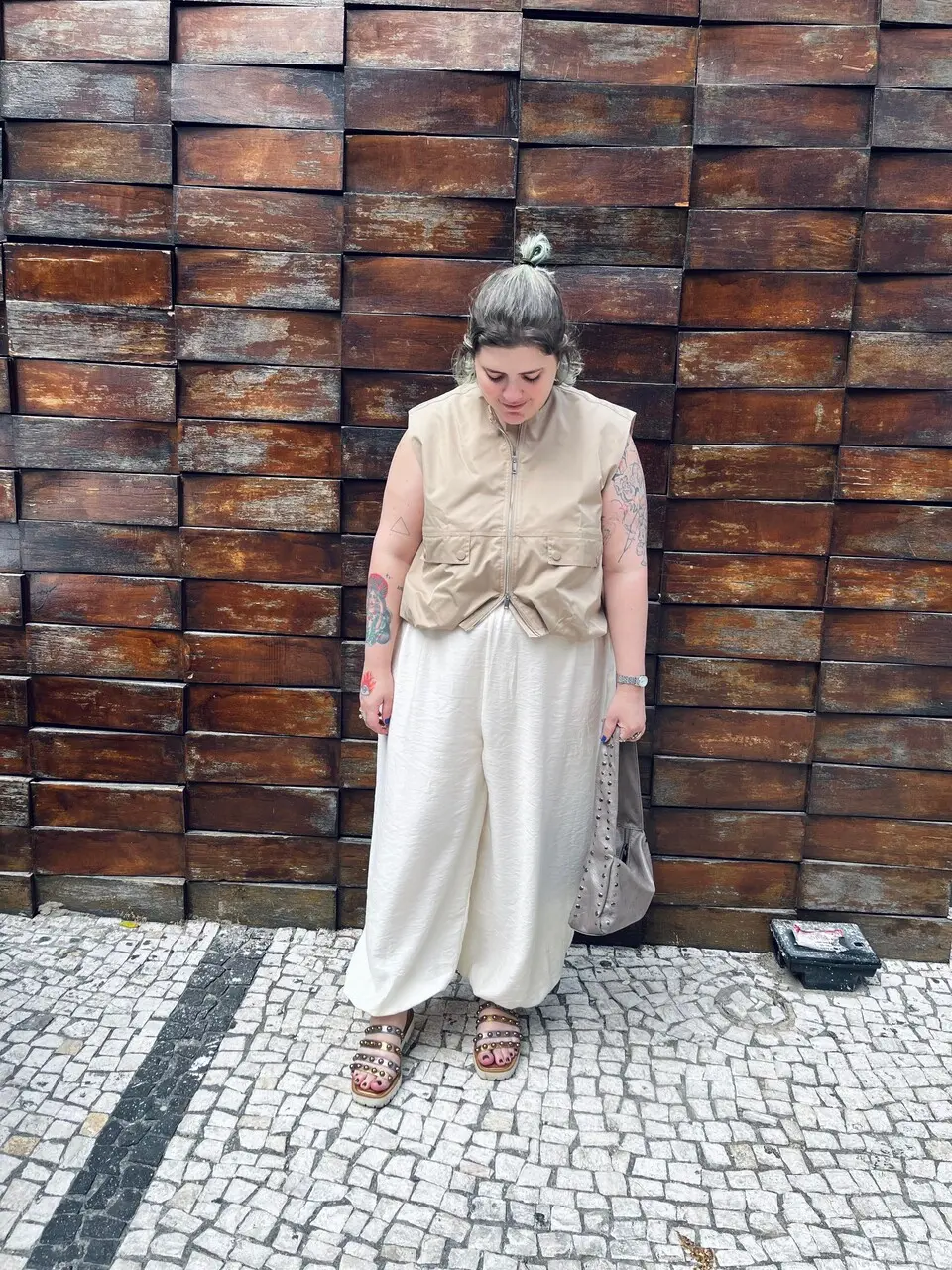 Uma mulher com cabelo descolorido preso em um coque alto está em pé na calçada de pedras portuguesas. Ela veste um colete bege utilitário sobre uma calça branca larga e solta, e sandálias plataforma douradas. Tatuagens são visíveis em seus braços. Ela segura uma bolsa marrom texturizada e usa um relógio prateado. O fundo é uma parede texturizada feita de blocos retangulares de madeira escura.