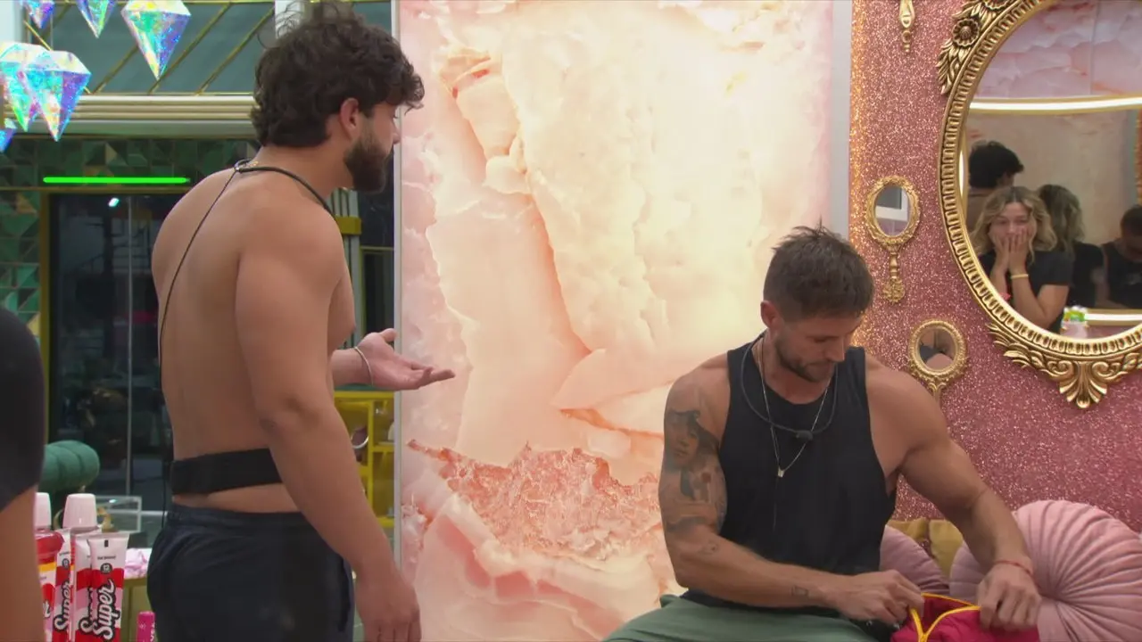 Dois participantes de reality show conversam: um homem sem camisa gesticula para outro sentado que arruma uma bolsa, enquanto uma mulher surpresa se reflete no espelho.