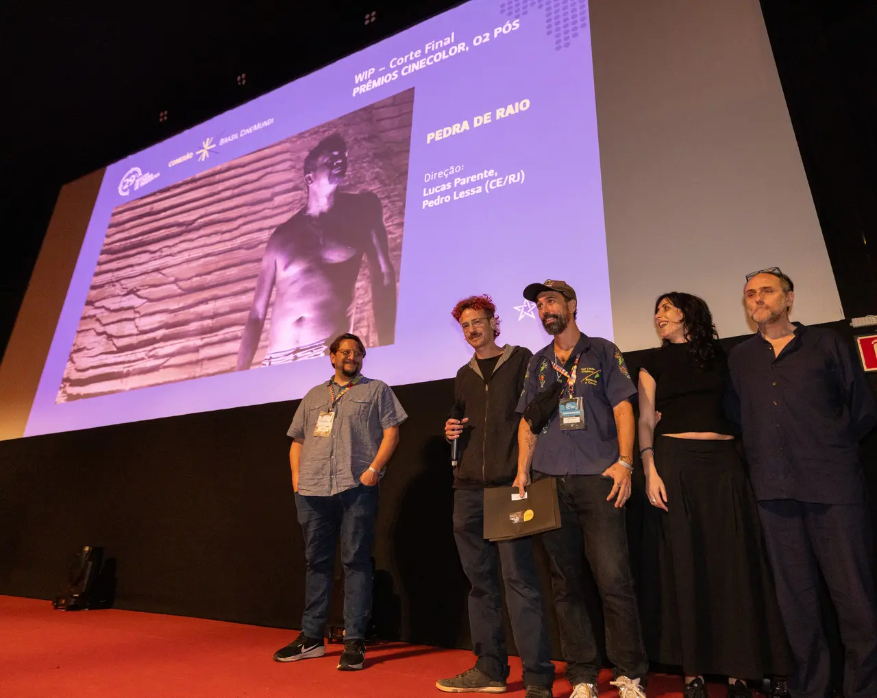 Cinco pessoas estão posicionadas no palco em frente a um grande telão que exibe informações sobre o filme 