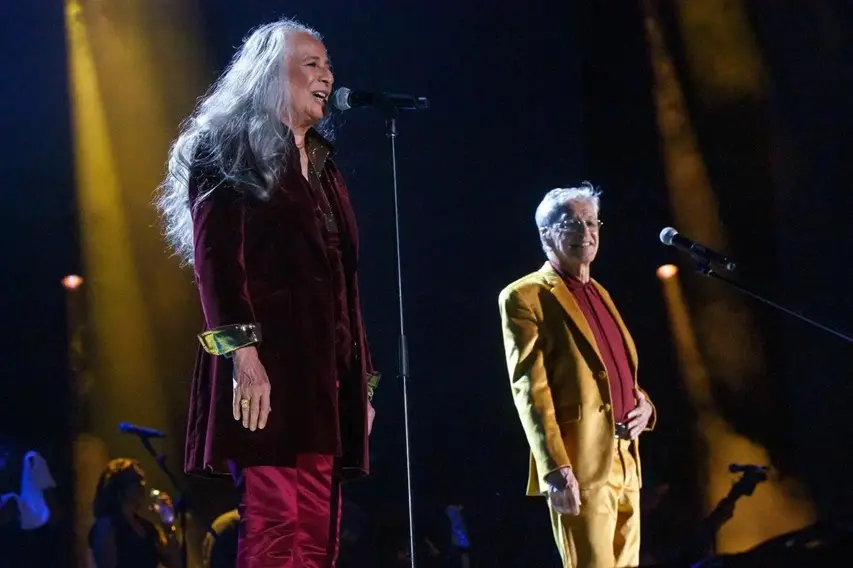 Maria Bethânia e Caetano Veloso cantam sorridentes no palco, iluminados por fortes feixes de luz dourada contra o fundo escuro. Bethânia veste um longo casaco de veludo vinho sobre calça acetinada, enquanto Caetano, ao fundo, usa um terno amarelo vibrante, compondo uma cena de grande elegância e cumplicidade musical.