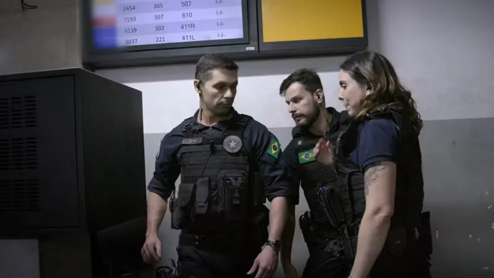 Três agentes da Polícia Federal, usando coletes táticos com a bandeira do Brasil, conversam em área interna que aparenta ser um aeroporto, com painéis de informação de voos ao fundo e equipamentos eletrônicos ao lado.