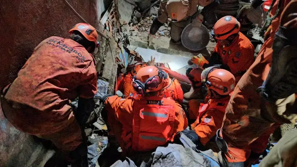 Equipe de resgate trabalhando em uma operação de busca após um desastre, com profissionais em uniformes laranja e capacetes, em um ambiente de destruição.