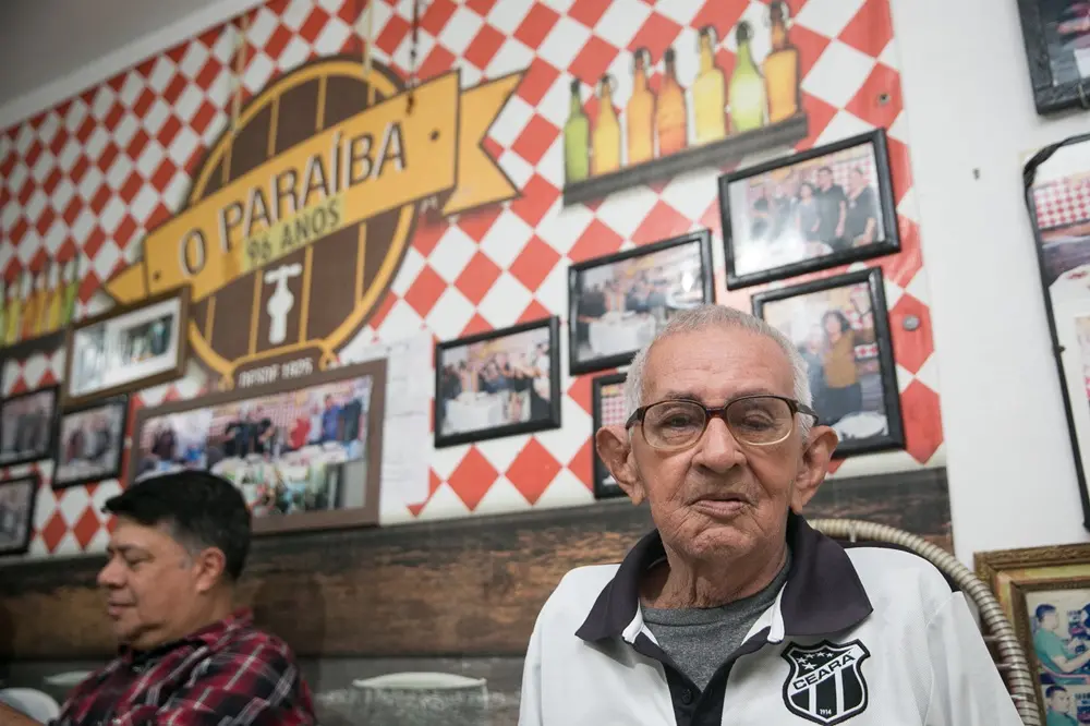 Na imagem, um homem idoso, de pele clara, com óculos de armação grossa e cabelos brancos curtos, aparece em primeiro plano olhando para a câmera. Ele veste uma camisa polo branca com golas pretas, que traz o escudo do Ceará Sporting Club no peito. Ao fundo, a parede é decorada com um padrão xadrez em vermelho e branco (estilo toalha de mesa de cantina). Nela, há um grande logotipo circular de madeira escrito 