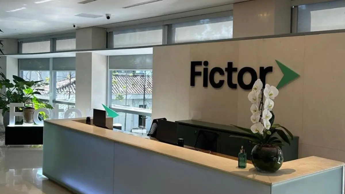 Recepção moderna de um escritório da empresa Fictor. Há um balcão amplo com tampo claro, duas estações de atendimento e um vaso com orquídeas brancas. Ao fundo, a parede exibe o logo “Fictor” com uma seta verde. Grandes janelas iluminam o ambiente com luz natural e há plantas decorativas ao redor.